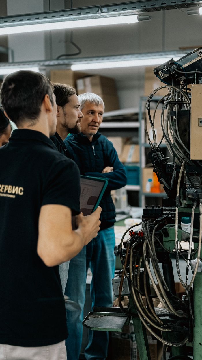 services-01 Three adults engaged in a discussion around complex machinery in a factory environment.